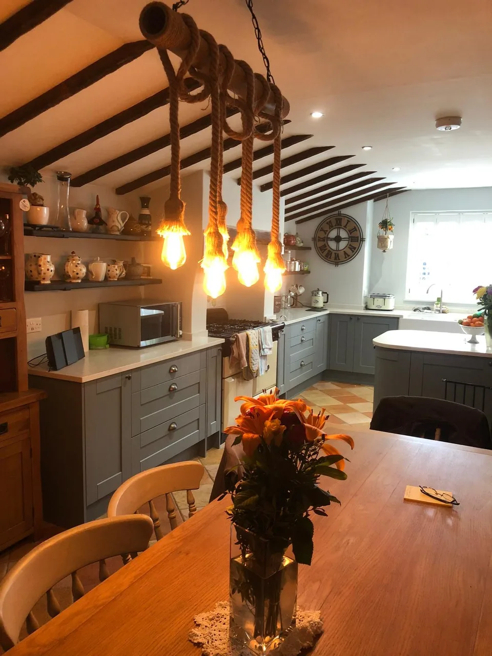 Kitchen and dining space with grey shaker cabinets, exposed ceiling beams and rope pendant lighting over a wooden table.